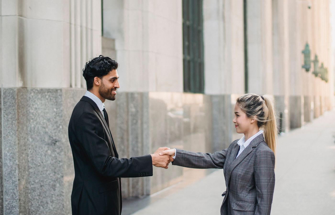 Two business professionals in suits exchanging a handshake outside a corporate building.