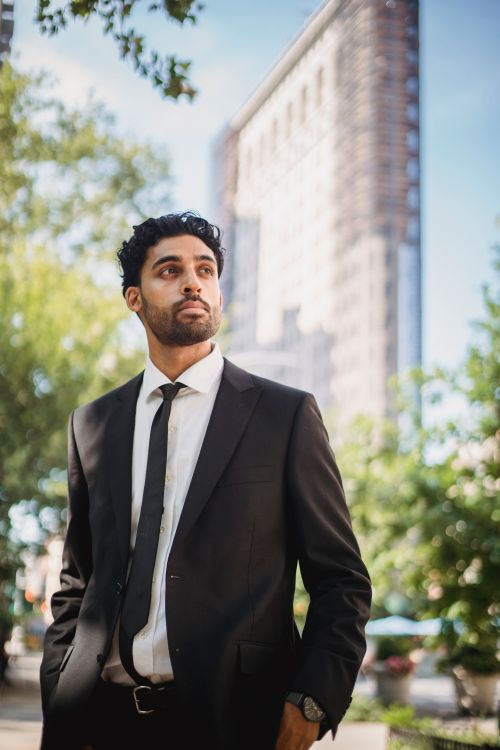 Free A confident young man in a black suit stands outside, looking afar against a highrise backdrop. Stock Photo