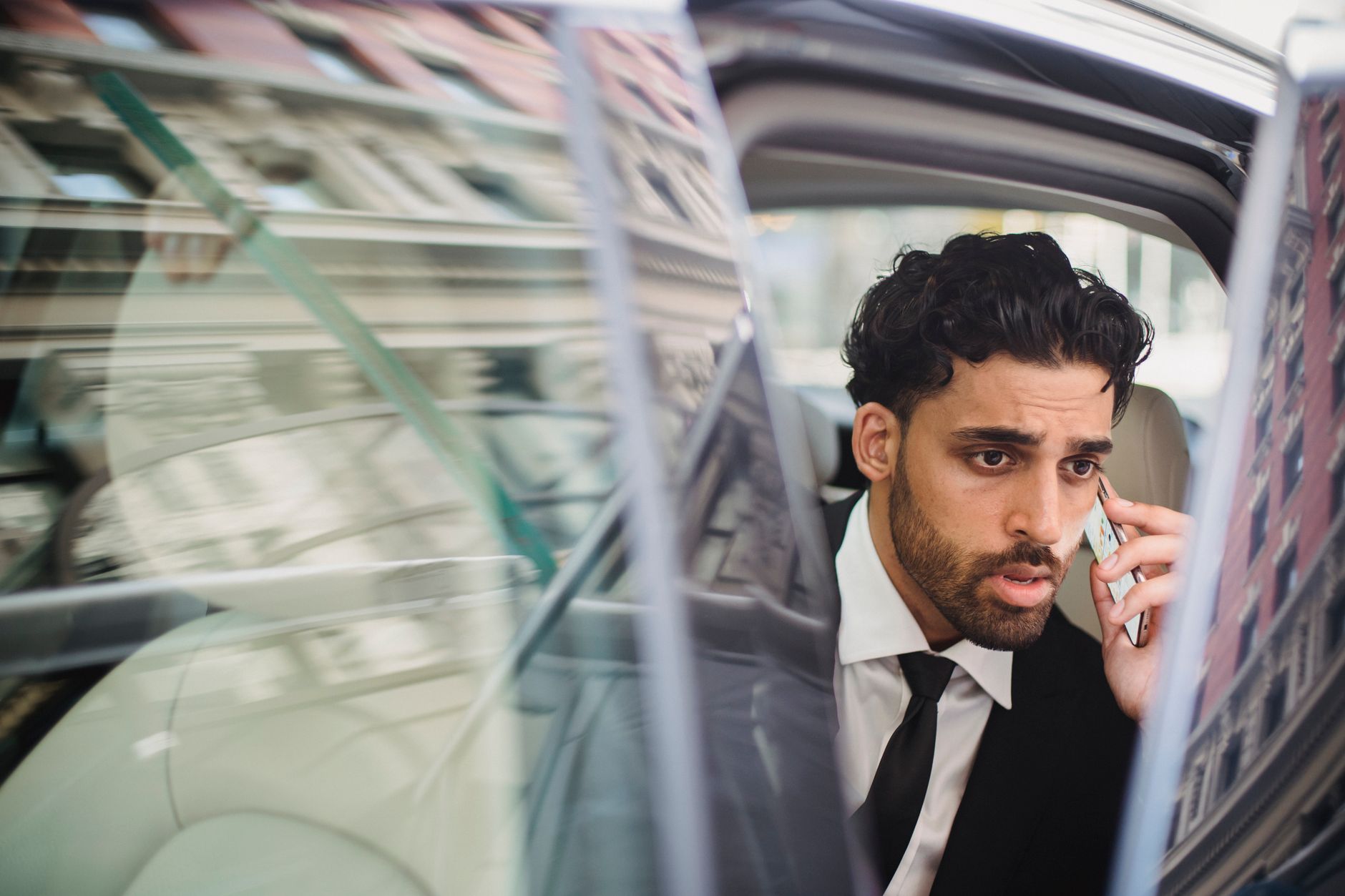 A professional man in a suit talking on his phone while seated in a car with an open door.