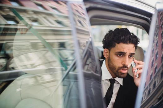 A professional man in a suit talking on his phone while seated in a car with an open door.