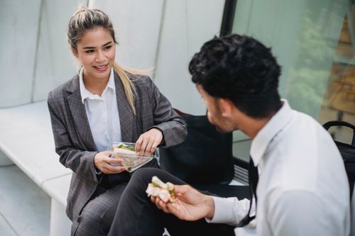Free Two business professionals in formal attire enjoy a lunch break outdoors, sharing sandwiches and conversation. Stock Photo