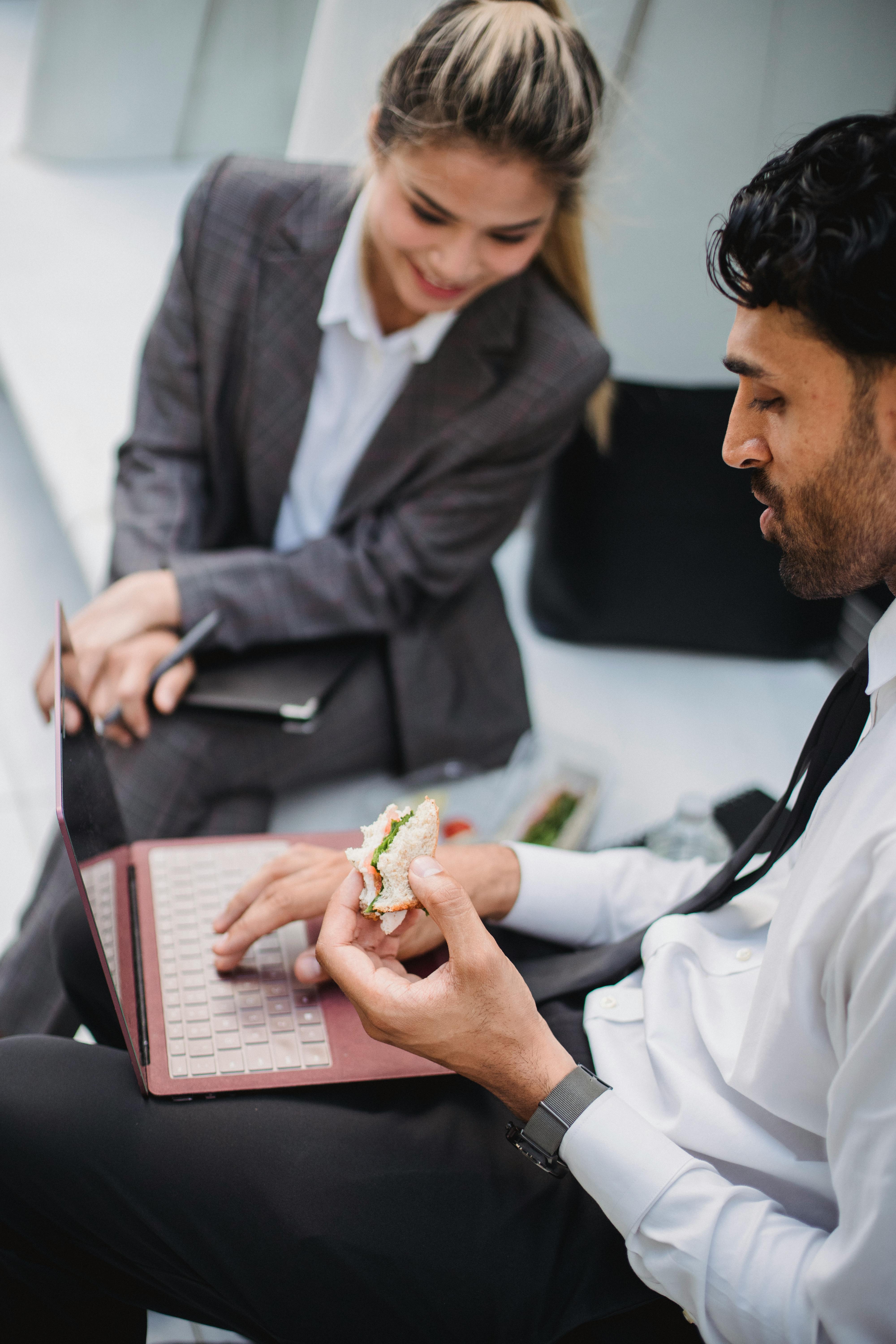 a bearded man sitting beside his colleague while using a laptop and eating