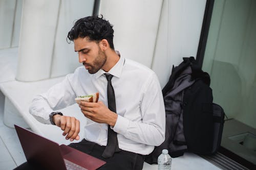 Free 
A Bearded Man in a Corporate Attire Looking at His Watch while Holding a Sandwich Stock Photo