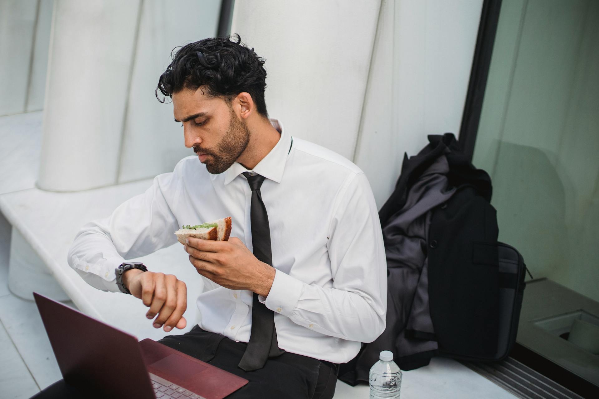 Businessman in formal attire eating a sandwich while checking his watch outdoors, multitasking during break.