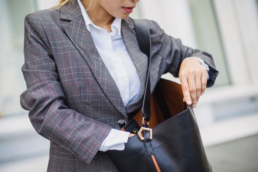 Close-up of a woman in a plaid blazer searching inside a leather bag outdoors.