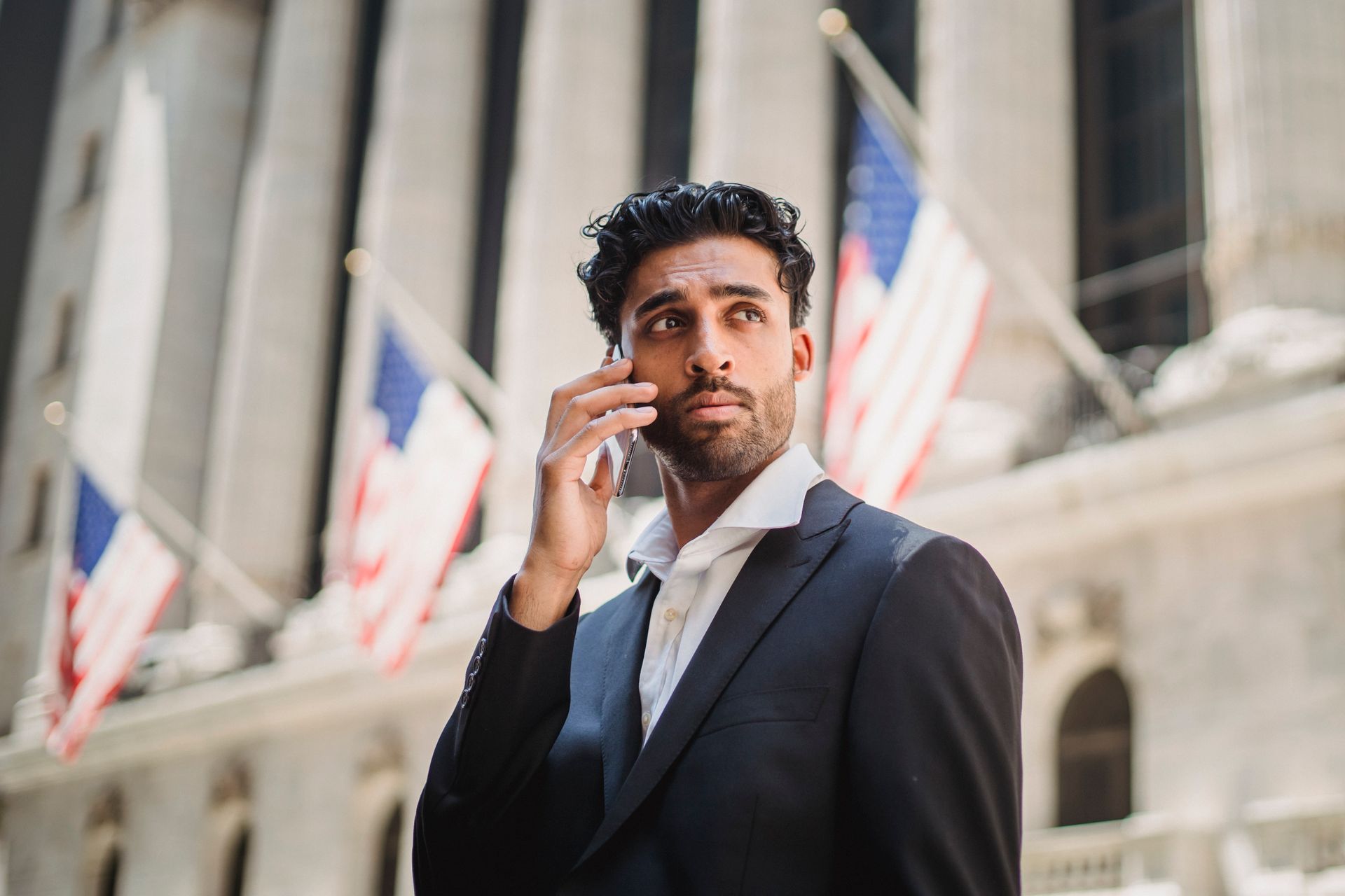 Businessman in black suit talking on mobile phone outside a historic building with flags.