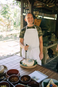 A cheerful male chef in a white apron prepares food in an outdoor kitchen, with a smile.