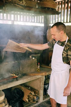 A man in an apron cooks using a wood stove in a rustic kitchen with smoke rising.