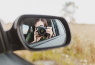 Reflection in car wing mirror of unrecognizable female traveler taking photo of rural field on camera during road trip in countryside
