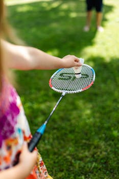 Close-up of child's hand holding a shuttlecock on a badminton racket outdoors.