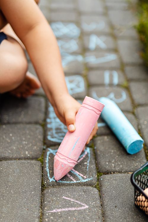 Free A child plays outdoors drawing with pink and blue chalk on a paved surface. Stock Photo