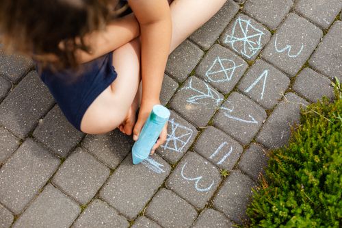 Free A child uses blue chalk to draw shapes and patterns on paved outdoor surface. Stock Photo