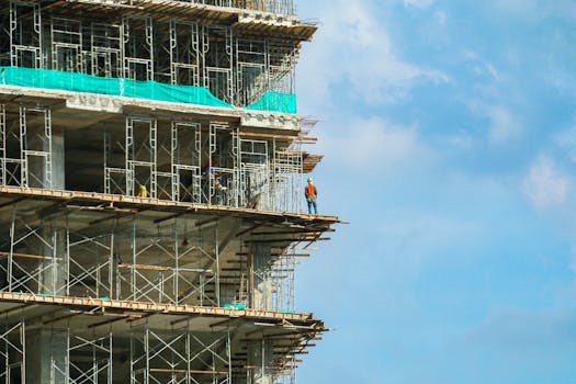 A construction worker stands on a scaffolding at a high-rise building under a blue sky.