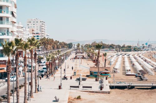 Free A bustling coastal promenade lined with palm trees, adjacent to a sunny beach with umbrellas and lounge chairs. Stock Photo