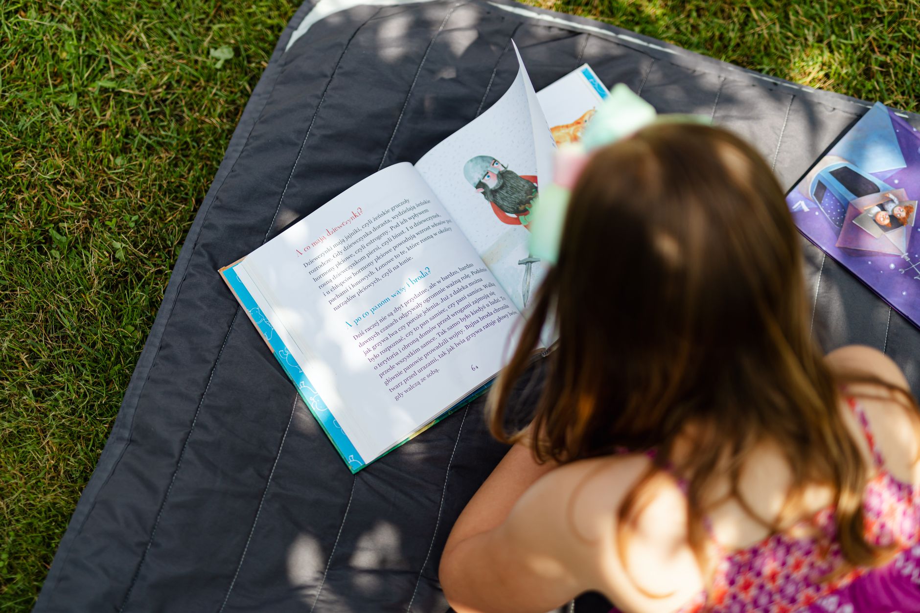 Girl with long hair reading book on blanket in the grass.