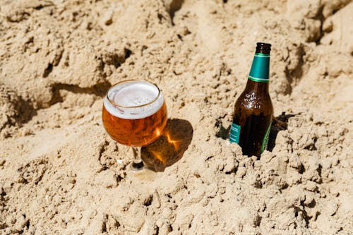 Beer Bottle Beside Clear Drinking Glass with Beer on Sand