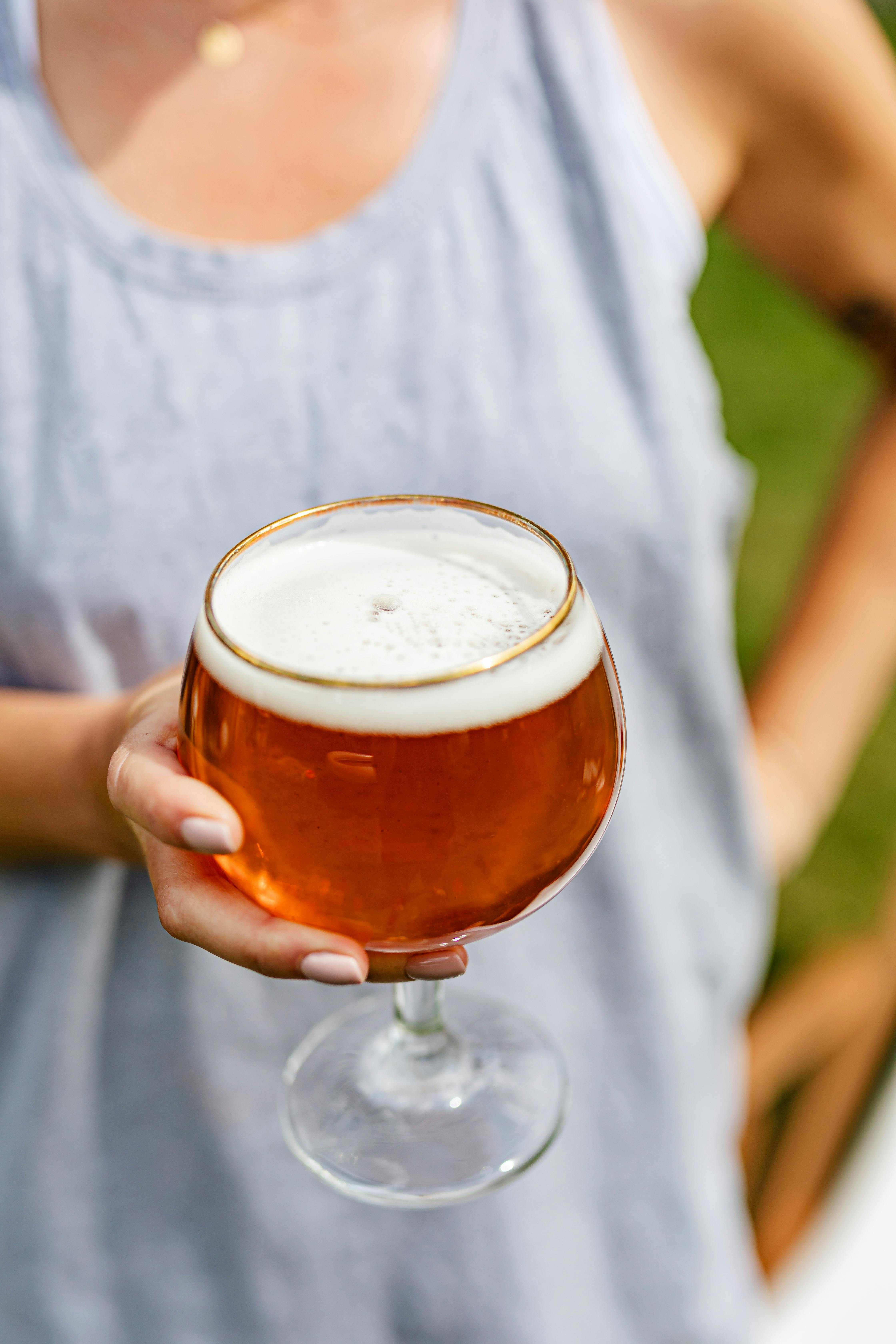 Close-up of a person holding a glass of frothy amber craft beer outdoors on a sunny day.