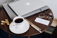 特写镜头对准木桌，上面放着笔记本电脑、咖啡、眼镜和文具，营造出舒适的工作空间氛围