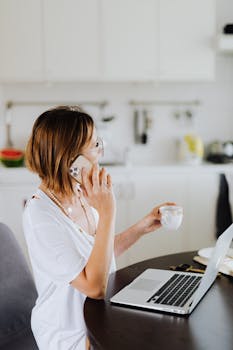 Woman multitasking with a smartphone and laptop while drinking coffee in a cozy home setting.
