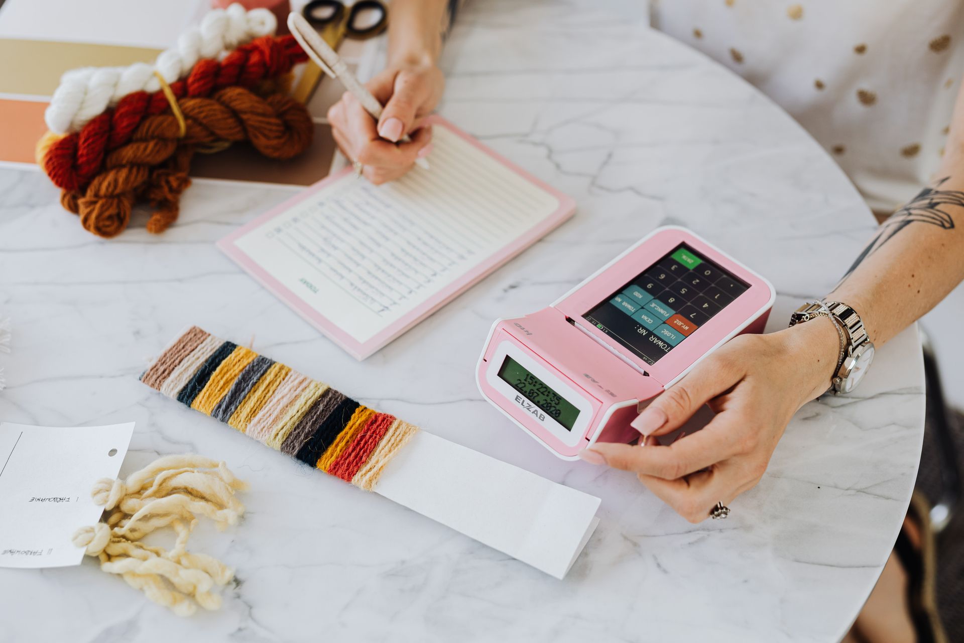 A person records details in a notebook while using a card reader for crafting supplies.