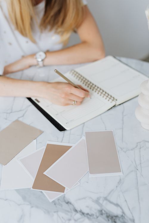 Free An adult woman writing in a notebook on a marble table with various neutral color swatches. Stock Photo