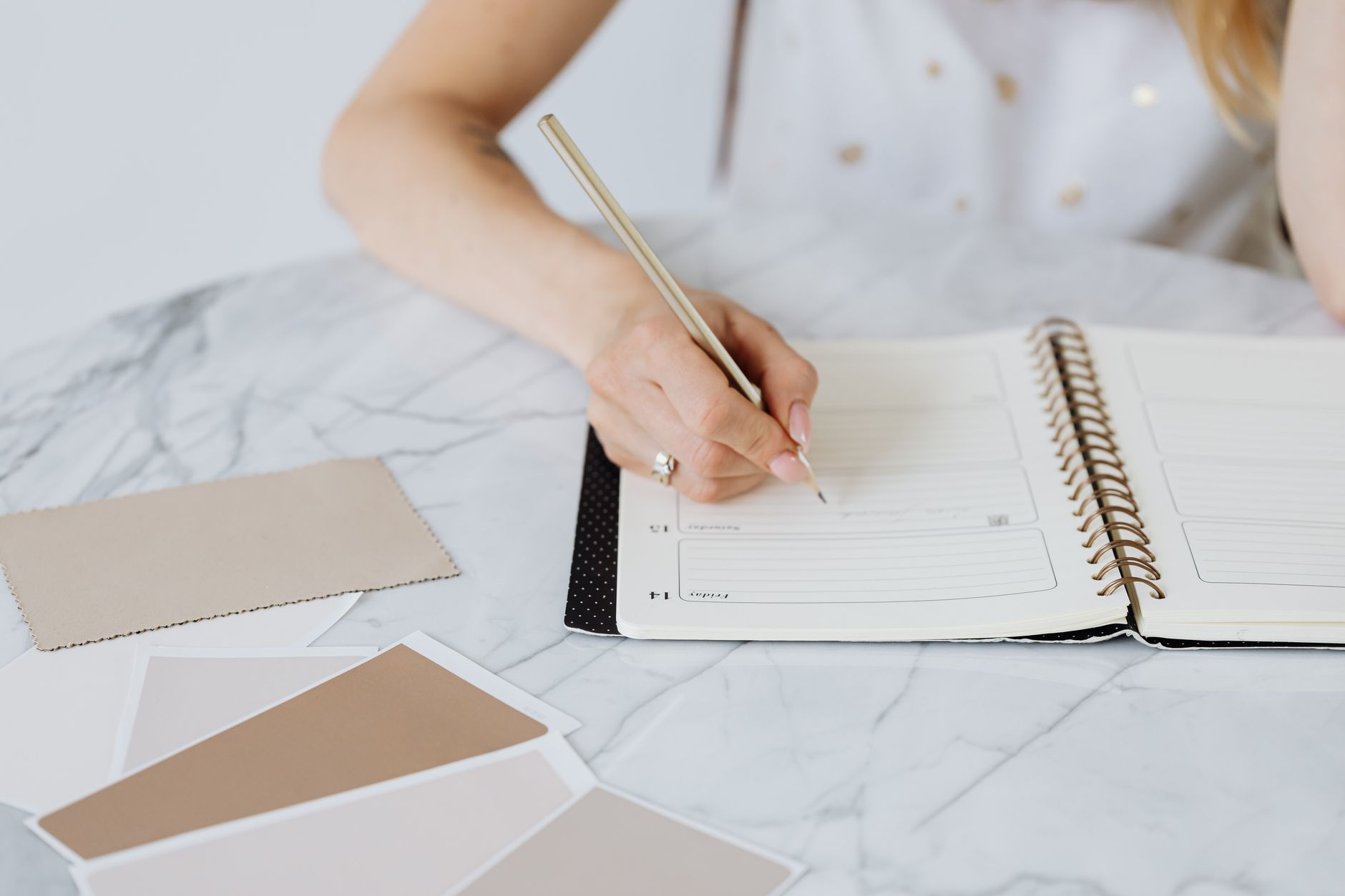 Close-up of a woman writing in a planner with color swatches on a marble table.