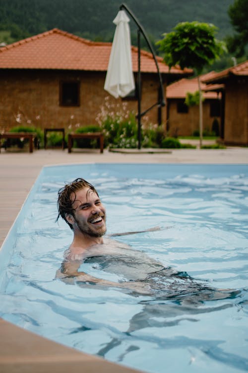 Happy man with long hair and beard swimming in pool near cottages against green hill