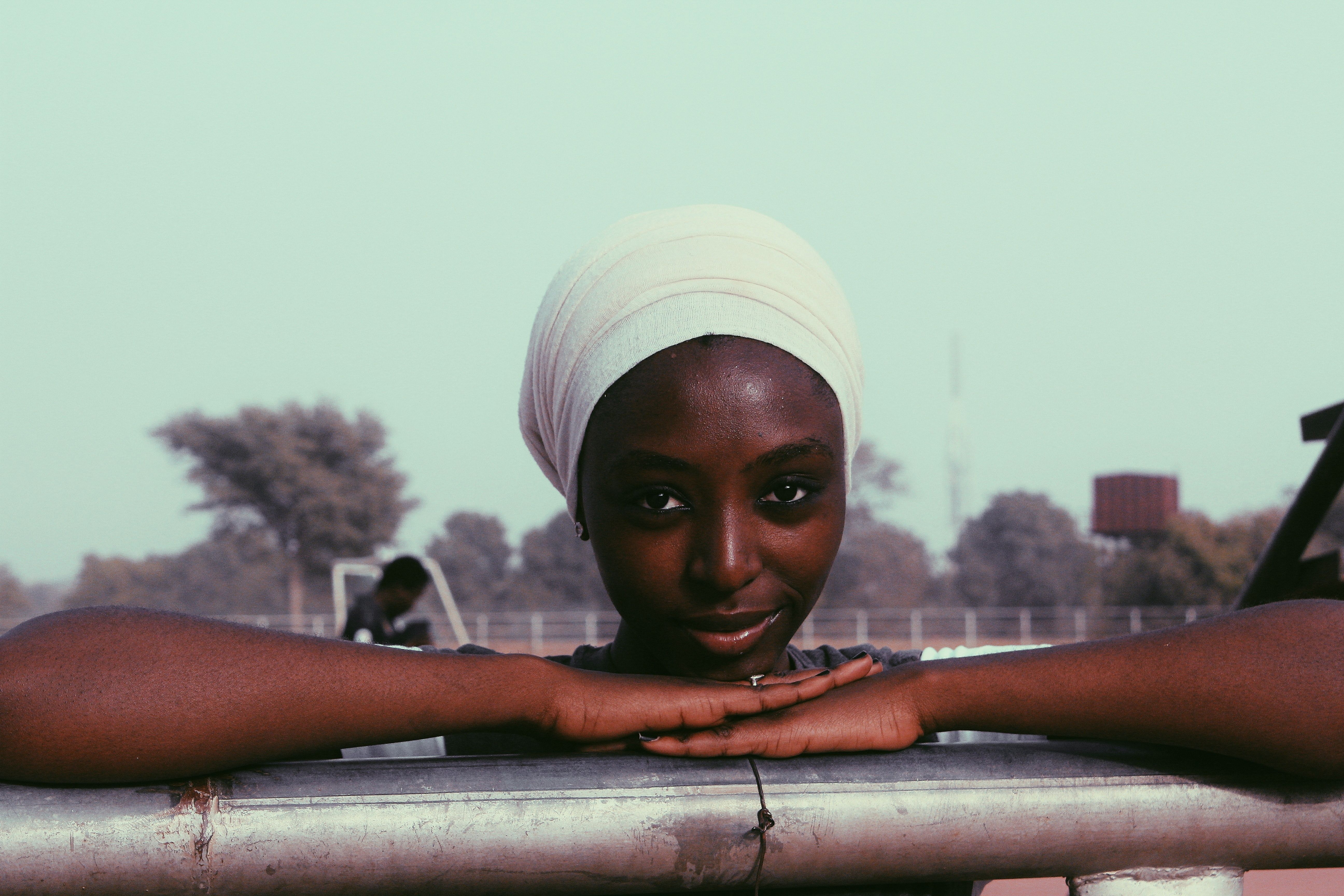 Confident young African American female in white turban leaning on metal fence smiling at camera on street