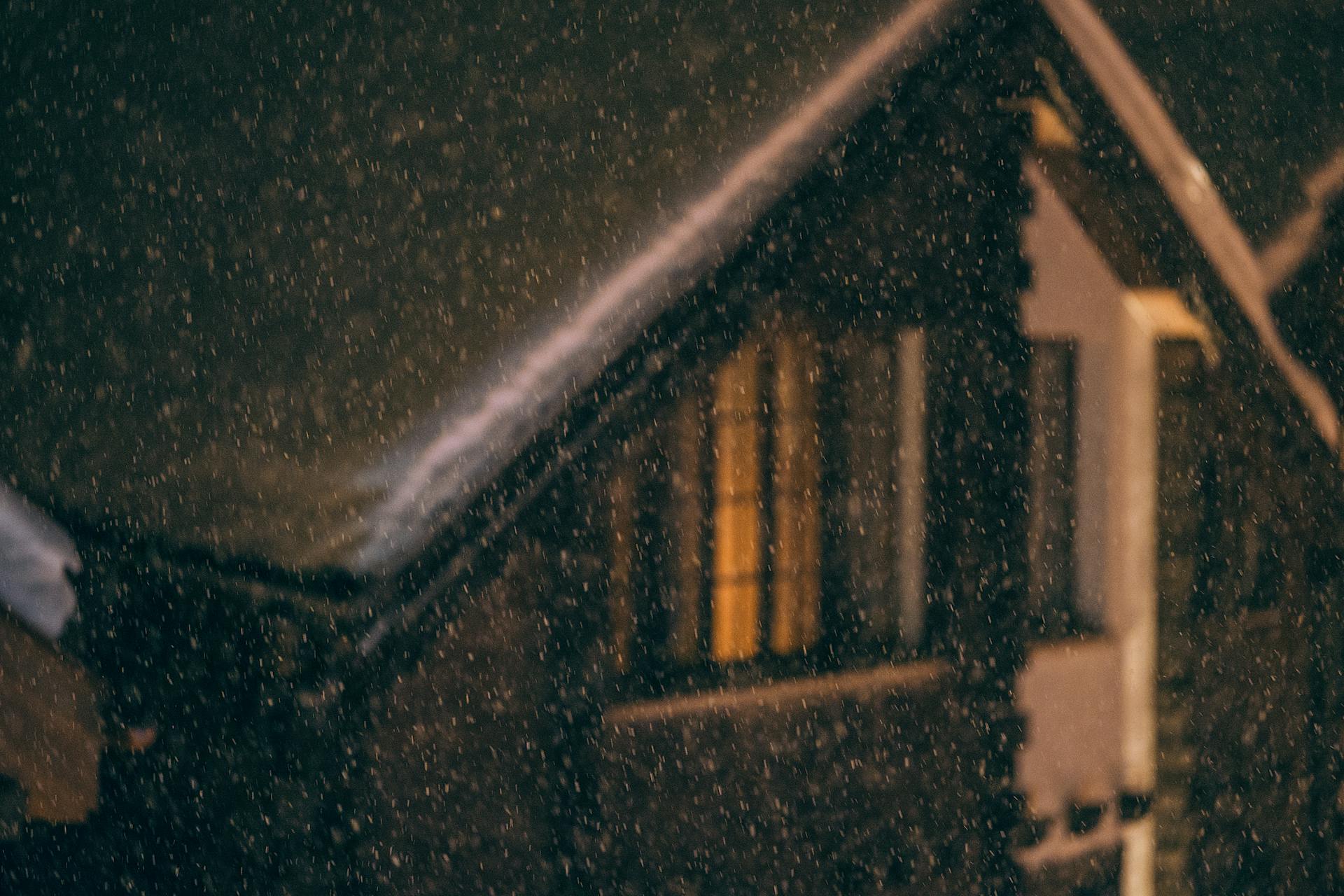 Exterior of small wooden cottage with snow on roof and burning electric lights in window during snowfall at cold winter night