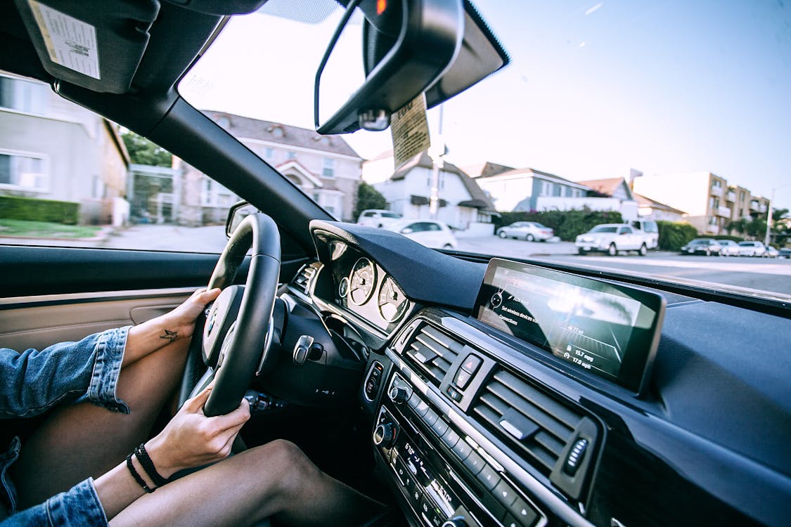 Free Side view of crop unrecognizable young female driver in denim jacket driving modern car on road near typical residential houses on sunny day Stock Photo