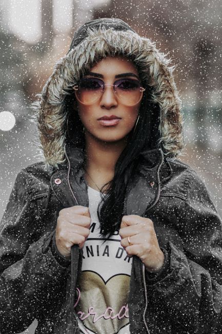 Portrait of a fashionable woman wearing sunglasses and a fur-lined jacket in a snowy setting.