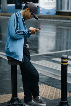 一名男子穿着休闲街头服饰，在下雨的街道上靠在护柱上使用手机
