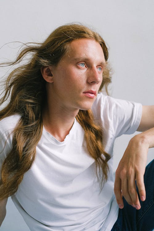 Free Young androgynous man with long hair sitting against white wall Stock Photo