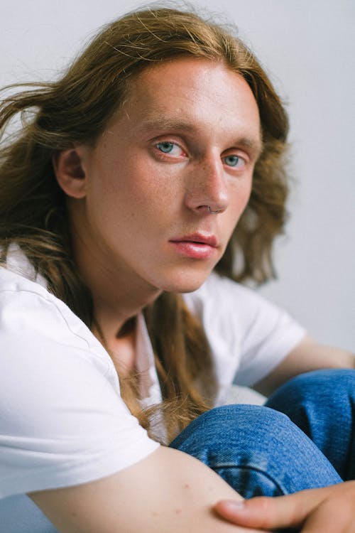 Unemotional young androgynous male with long ginger hair embracing knees and looking at camera while sitting against white wall in studio