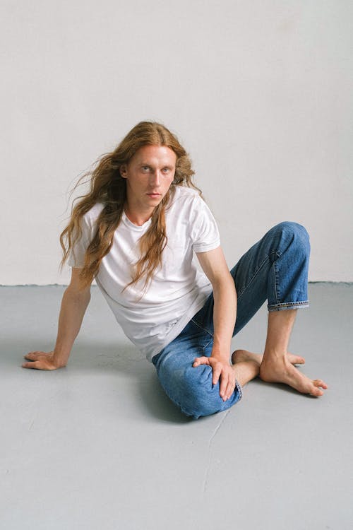 Full length young androgynous male model with long ginger hair sitting on floor in light studio and looking at camera