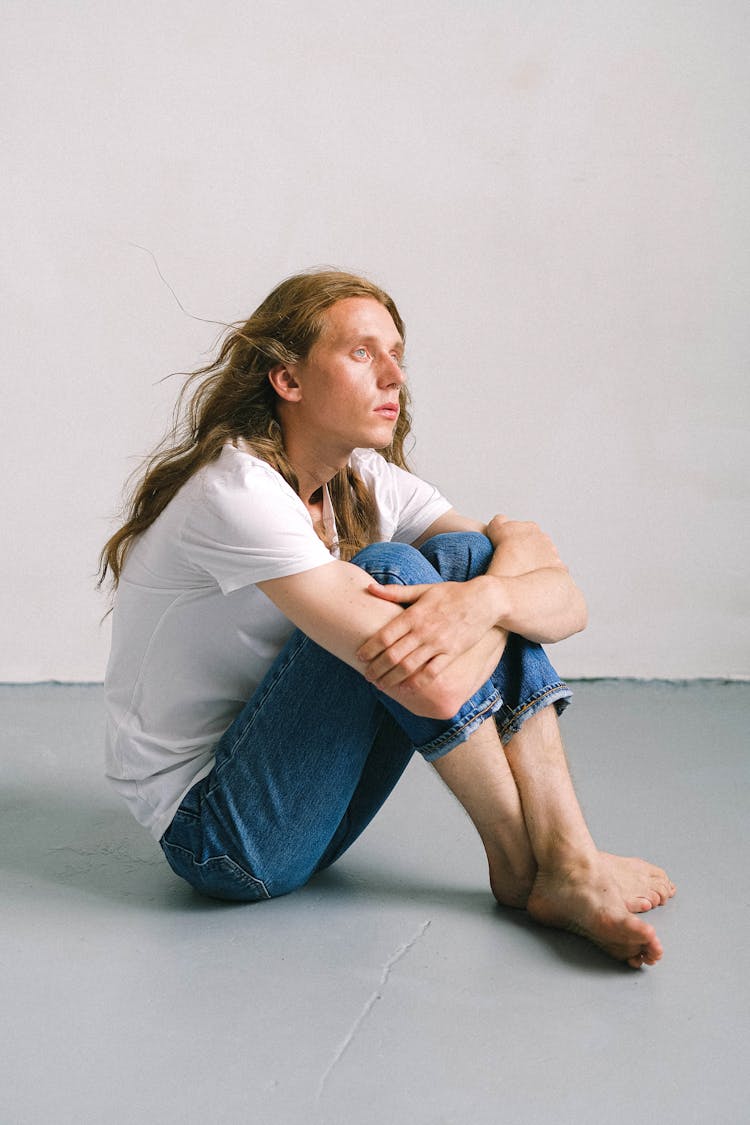 Wistful Androgynous Man Embracing Knees In Light Studio