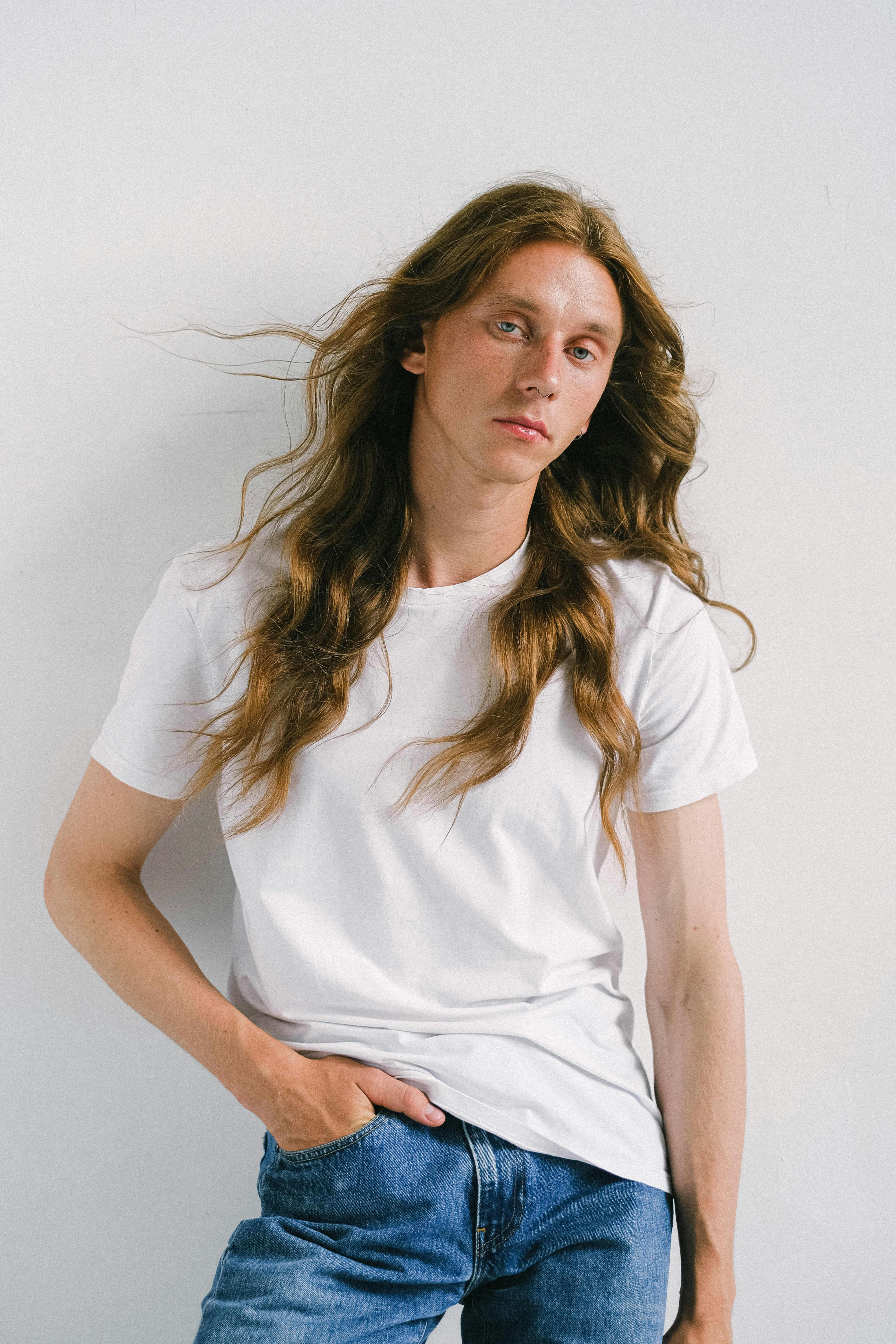 Calm androgynous male model with long hair standing with hand in pocket against white wall and looking at camera