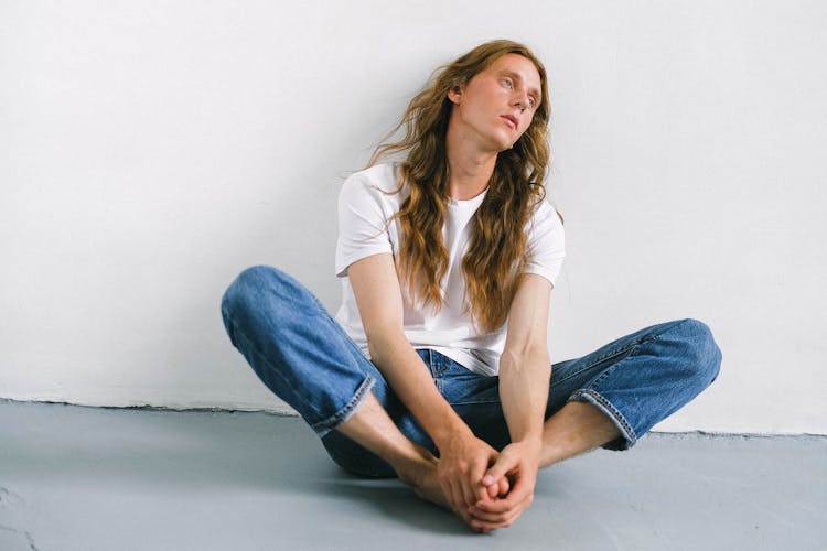 Pensive Androgynous Man Sitting On Floor With Legs Apart