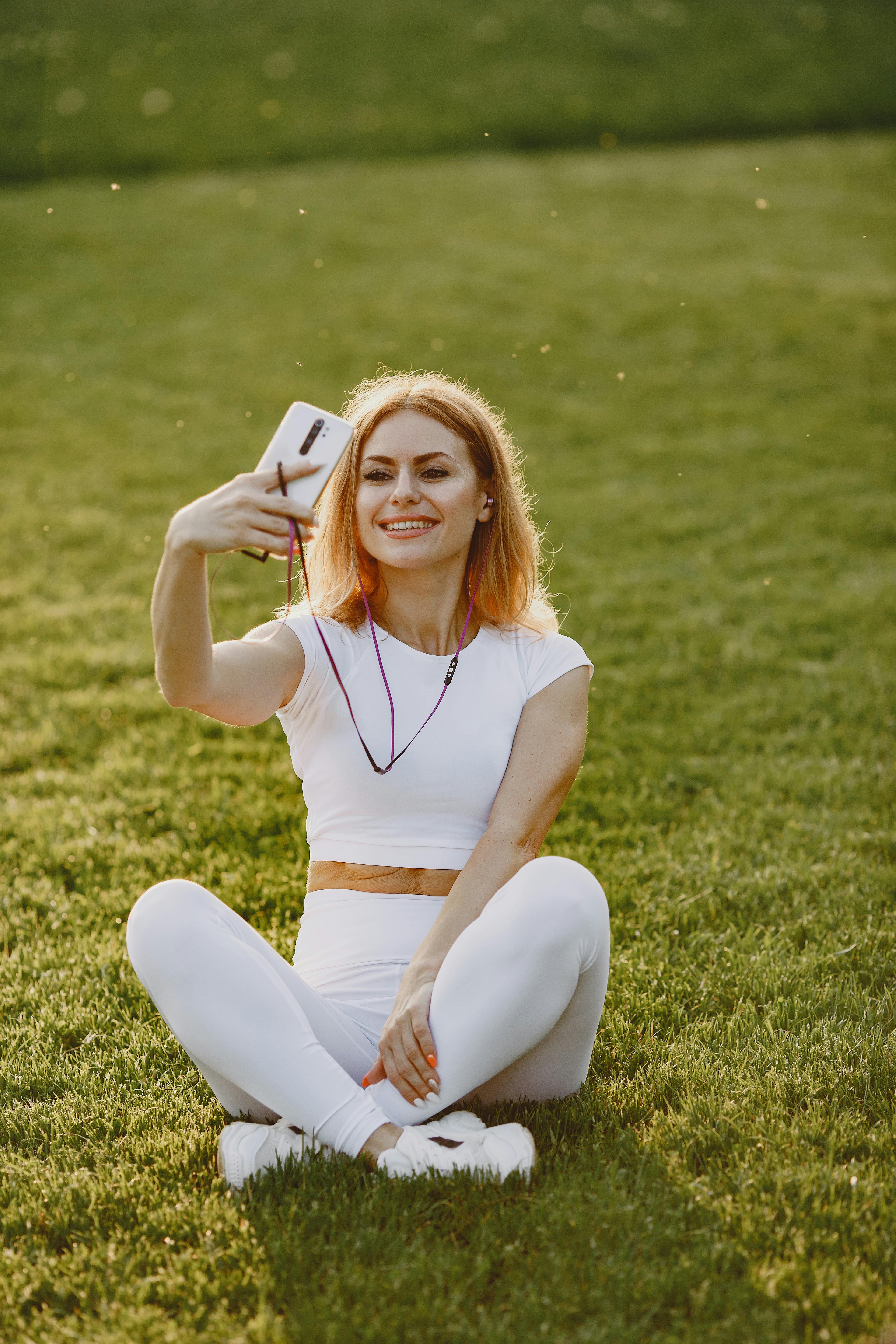 woman in white activewear sitting on the grass field while taking photo of herself