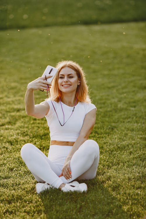Free Woman in White Tank Top and White Pants Sitting on Green Grass Field Stock Photo