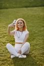 Woman in White Tank Top and White Pants Sitting on Green Grass Field