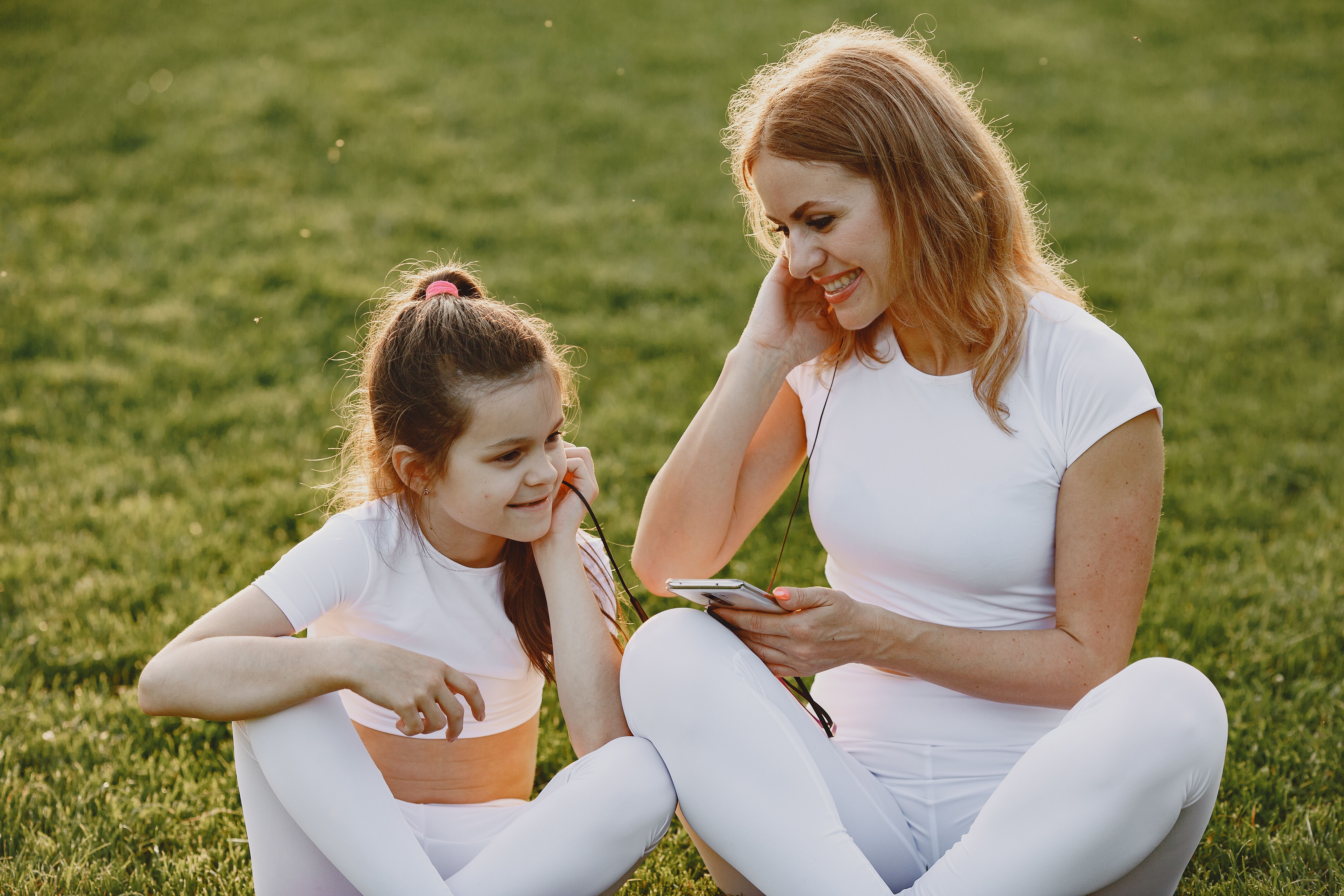 A mother and daughter sitting on grass, sharing headphones and enjoying music outdoors.