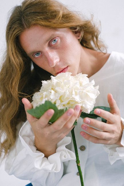 Dreamy young guy with long wavy hair in feminine outfit holding delicate white bigleaf hydrangea flower in hand and looking at camera