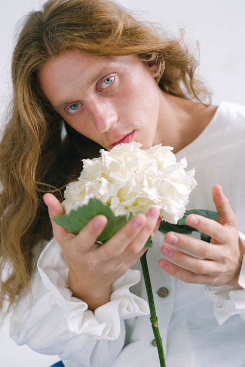 Free Dreamy young guy with long wavy hair in feminine outfit holding delicate white bigleaf hydrangea flower in hand and looking at camera Stock Photo