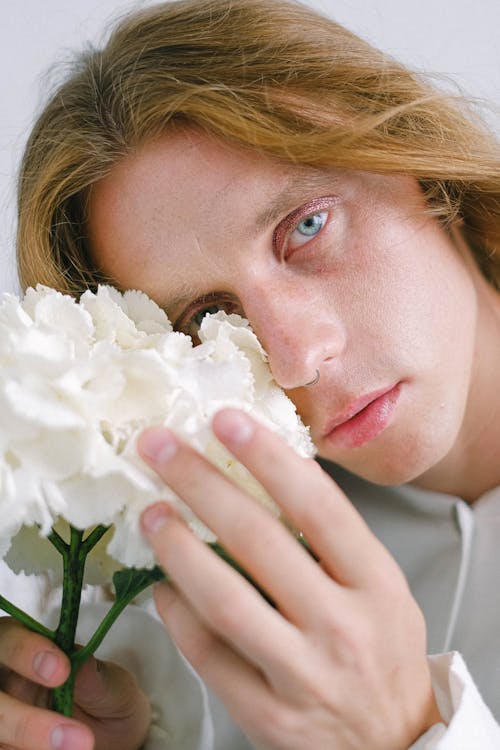 Free Pensive young androgynous male covering eye with flowers Stock Photo