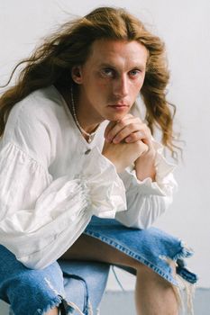 一位留着长波浪头发、穿着白衬衫和牛仔裙的古怪年轻男子，坐在椅子上看着镜头