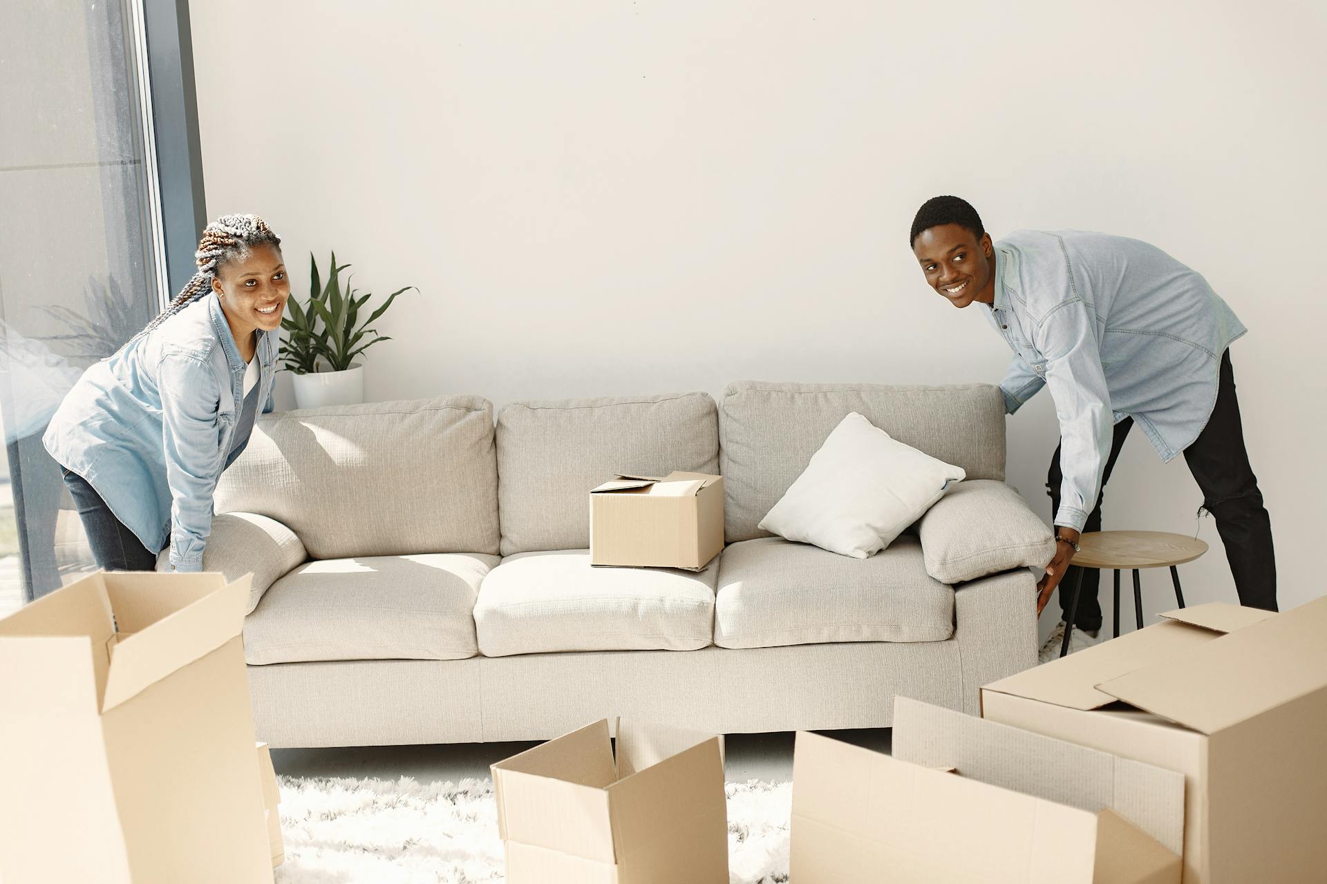 A cheerful couple moving furniture in a well-lit modern living room with cardboard boxes.