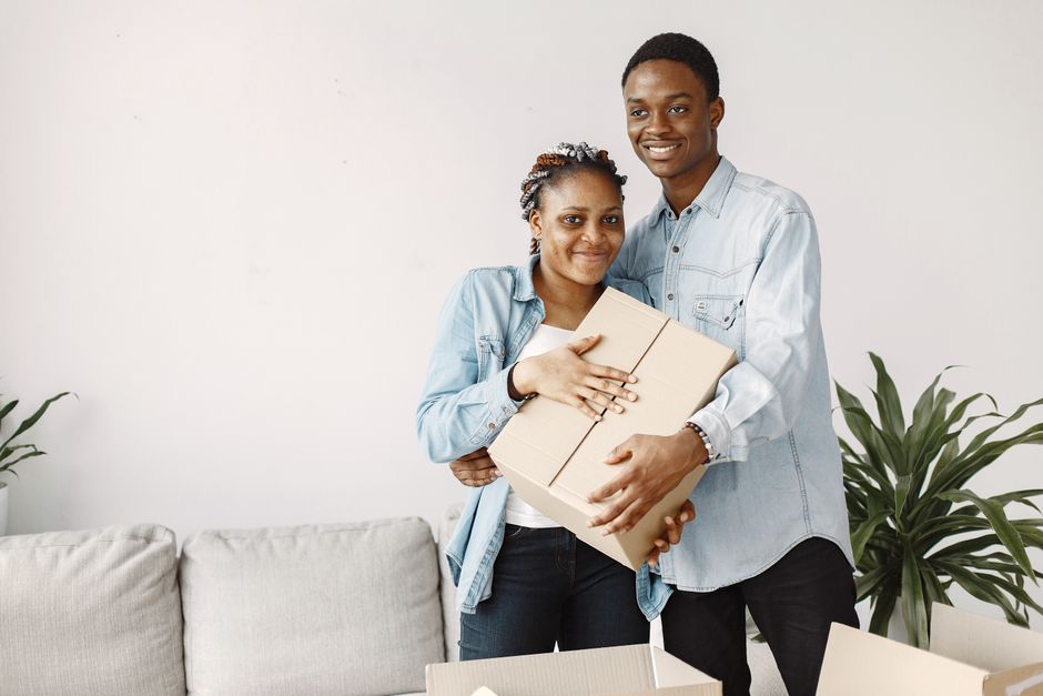 Group of young Black Nigerian friends in a Birmingham living room unpacking a shared bulk grocery order, bags of yam and plantain stacked on the floor, warm evening indoor light, photorealistic