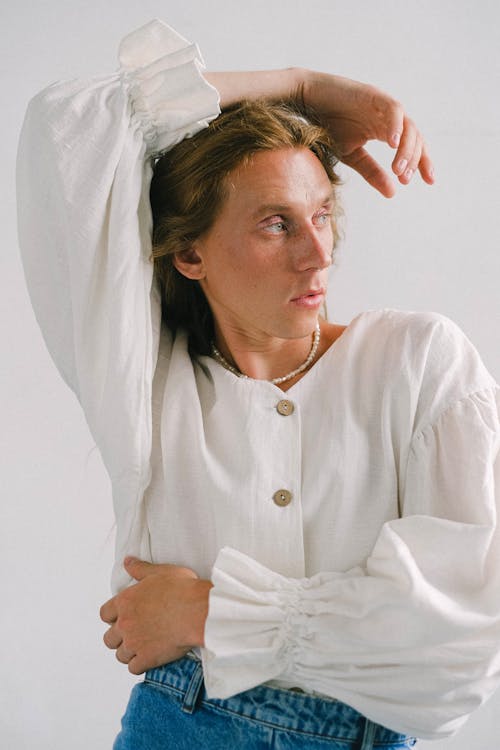 Free Fashionable young androgynous man in white blouse looking away with hand on head while standing in studio Stock Photo