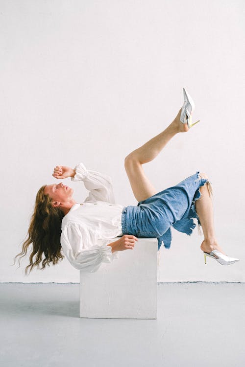 Side view of eccentric gender fluid young guy with long wavy hair in denim skirt and white blouse lying on cube with raised legs